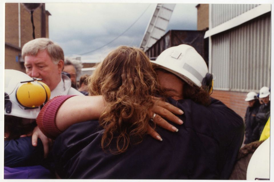 Brenda Procter, wearing a hard hat, embraces a woman seen from behind, moments after the Trentham 3 emerged from the pit. Several other people are visible in the background.